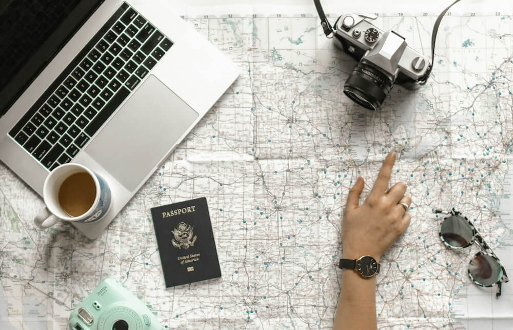 person-pointing-countries-on-the-map-which-is-on-the-table-where-there-are-passport-laptop-camera-and-coffee